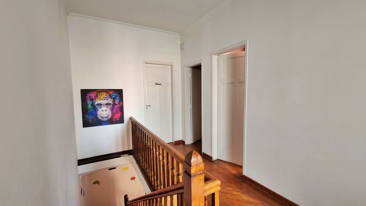 A well-lit hallway features a wooden staircase leading to various rooms. A vibrant artwork of a monkey hangs on the white wall, adding a pop of color. Light reflects off the smooth wooden floor, creating a warm ambiance.