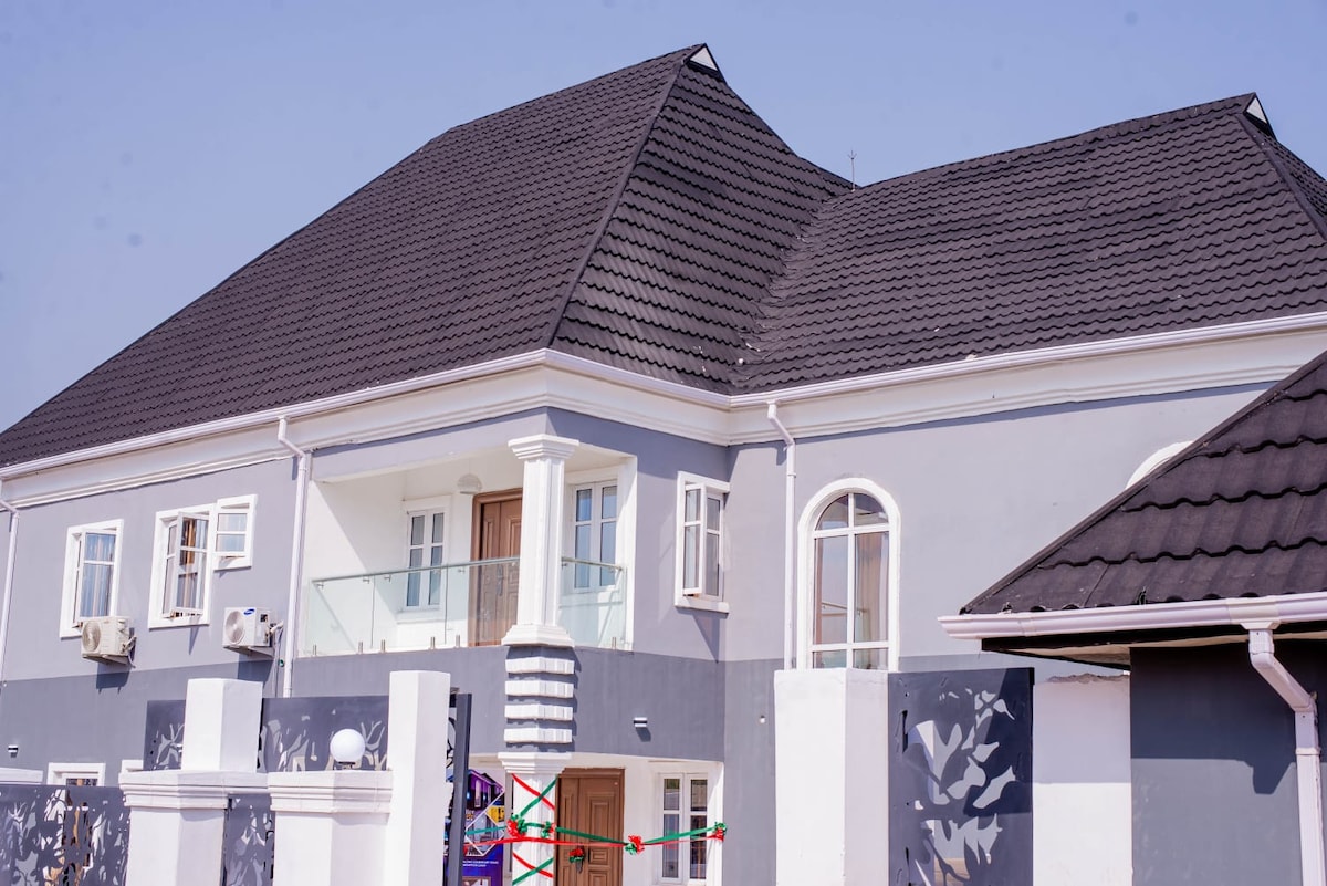 A two-story luxury home features a modern architectural design, with a grey exterior and white trim. A covered balcony is visible above the main entrance, framed by large windows. The entrance is marked by double doors, and decorative elements enhance the property’s inviting look.