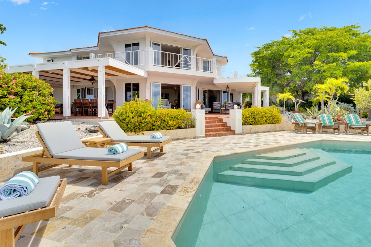 A spacious outdoor area features a swimming pool with steps leading in, surrounded by comfortable lounge chairs. The villa's exterior is accentuated by a wraparound porch and vibrant tropical landscaping, providing a serene backdrop for relaxation and outdoor gatherings.