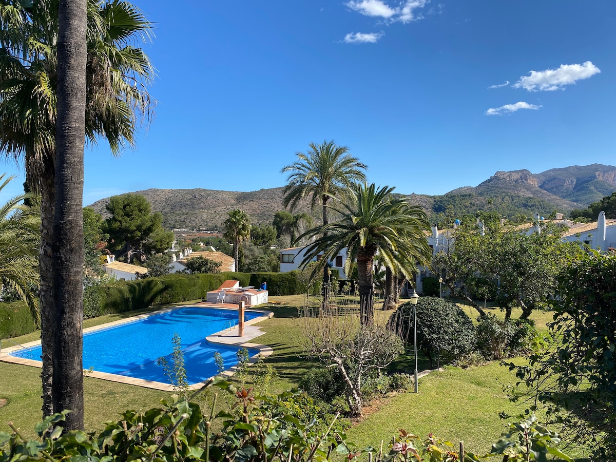 A tranquil outdoor space is depicted, featuring a vibrant pool surrounded by palm trees and lush greenery. Majestic mountains rise in the background, while the sunny sky enhances the peaceful atmosphere of the landscape.