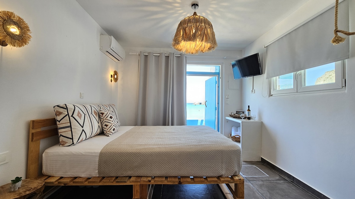 A bright and airy room features a spacious bed adorned with decorative pillows. Natural light enters through a partially open door, revealing a view of the sea. A television is mounted on the wall, and air conditioning is available for comfort.