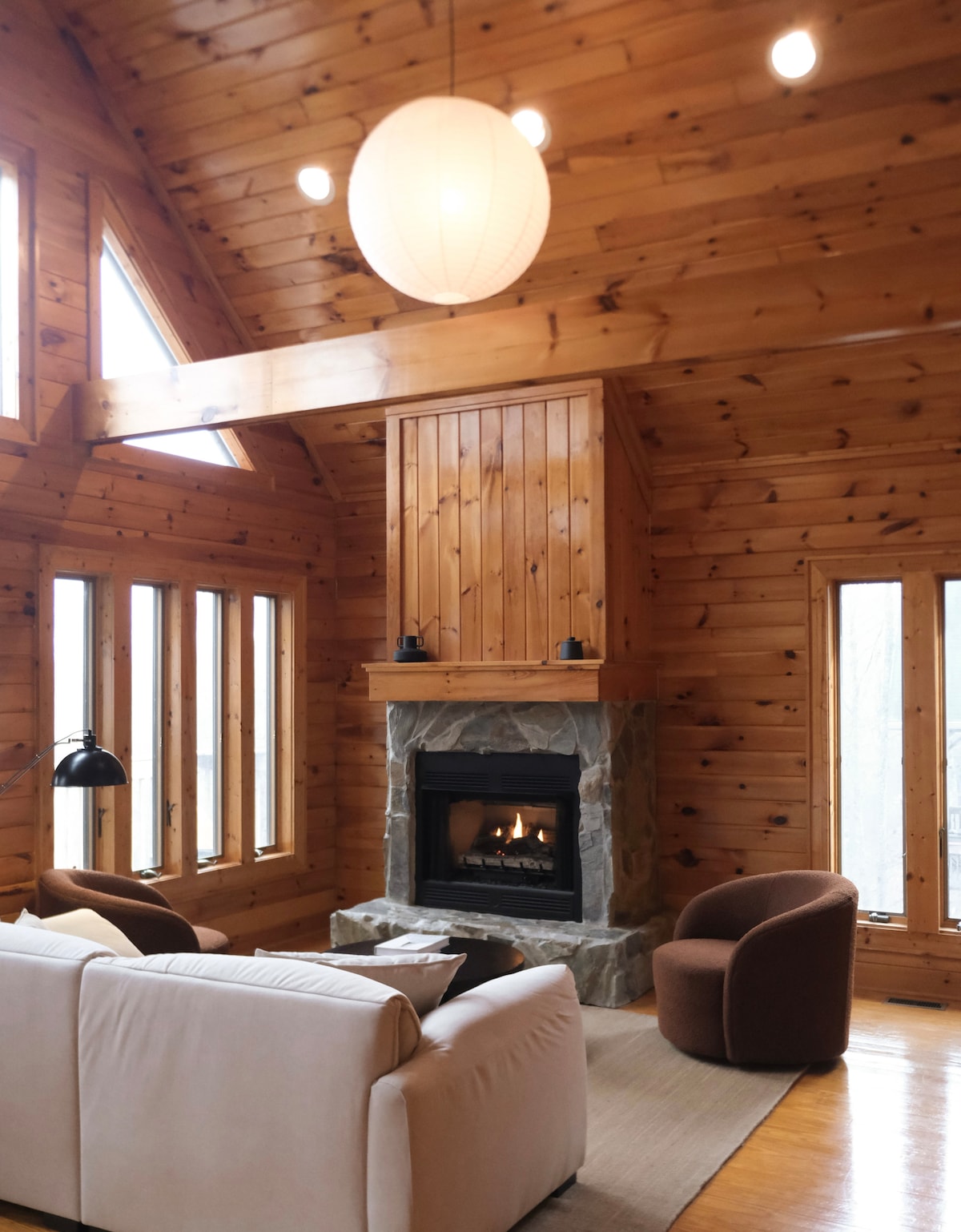 The inviting living area features warm wooden walls and vaulted ceilings, complemented by large windows allowing natural light. A modern fireplace is centered against a stone backdrop, surrounded by comfortable seating including a white sofa and two brown accent chairs.