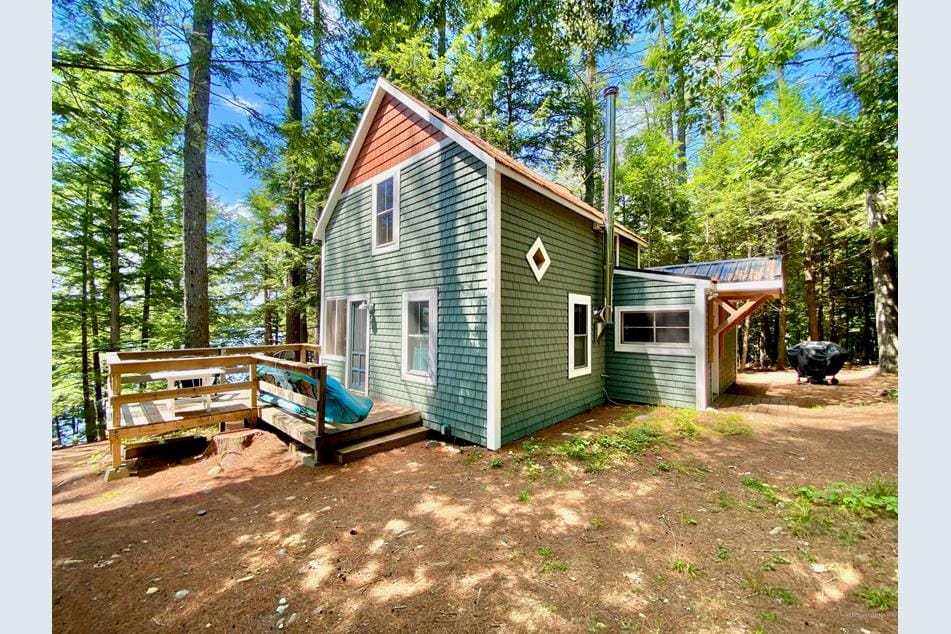 The exterior of a renovated camp is shown, featuring a green façade with a rustic charm. Large windows and multi-colored roof accents add character. A wooden deck with a kayak rests nearby, while tall pine trees create a shaded, natural atmosphere around the cozy structure.