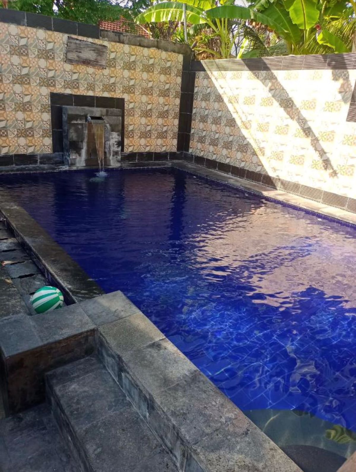 A serene swimming pool with clear blue water is framed by intricately tiled walls. A gentle water feature cascades into the pool, while a patterned ball rests near the edge. Lush greenery is visible in the surrounding area, enhancing the peaceful atmosphere.