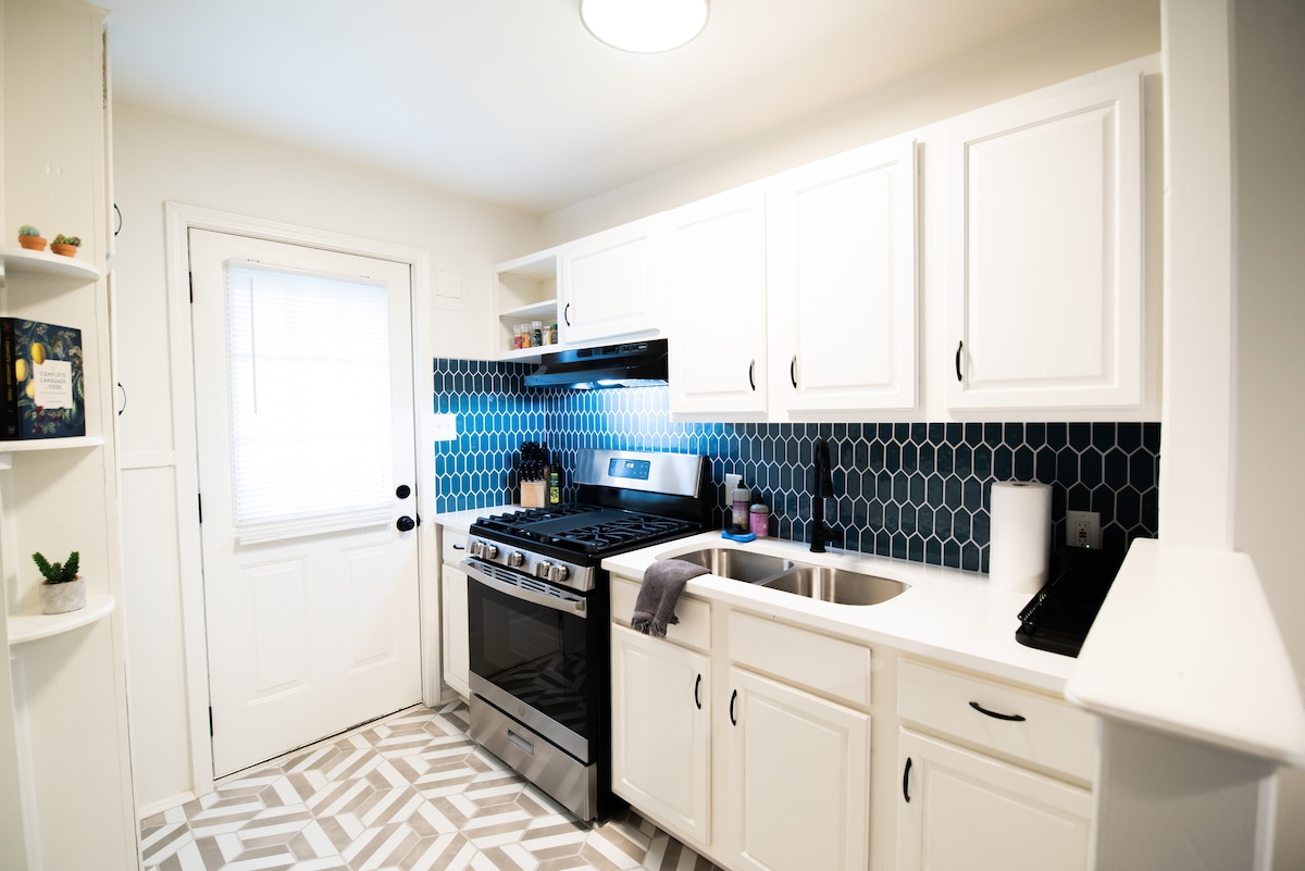 The kitchen features white cabinets contrasted by a hexagonal blue backsplash. A modern stainless steel stove is positioned next to a double sink. Natural light enters through a nearby window, illuminating the stylish patterned flooring.