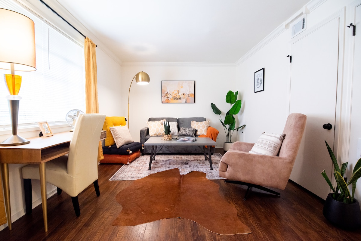 The living room features a mix of contemporary furniture, including a gray couch and a light-colored armchair. A wooden table is positioned alongside a floor lamp, and a large plant adds a touch of greenery. A colorful area rug grounds the space, complementing the warm wood flooring.