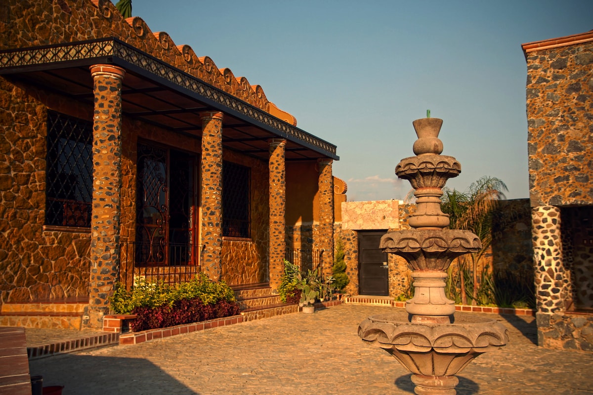 A charming exterior view of a stone structure showcases elegant columns and a tiered fountain surrounded by lush greenery. Paved stone pathways lead to the entrance, while the warm light illuminates the building's unique architecture.