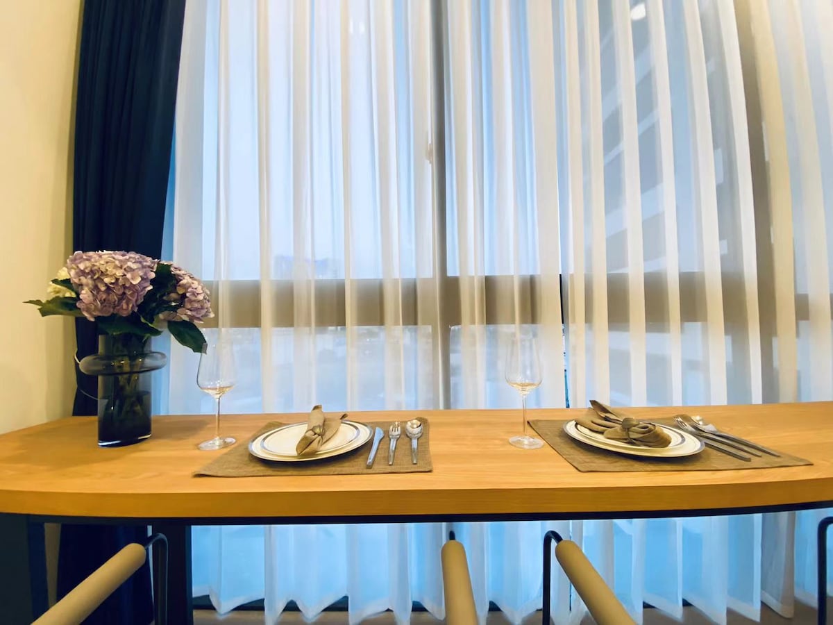 A dining table is set with elegant dishware and glassware, accompanied by a decorative vase holding fresh flowers. Light filtering through sheer curtains creates a soft ambiance, enhancing the inviting atmosphere of the space.