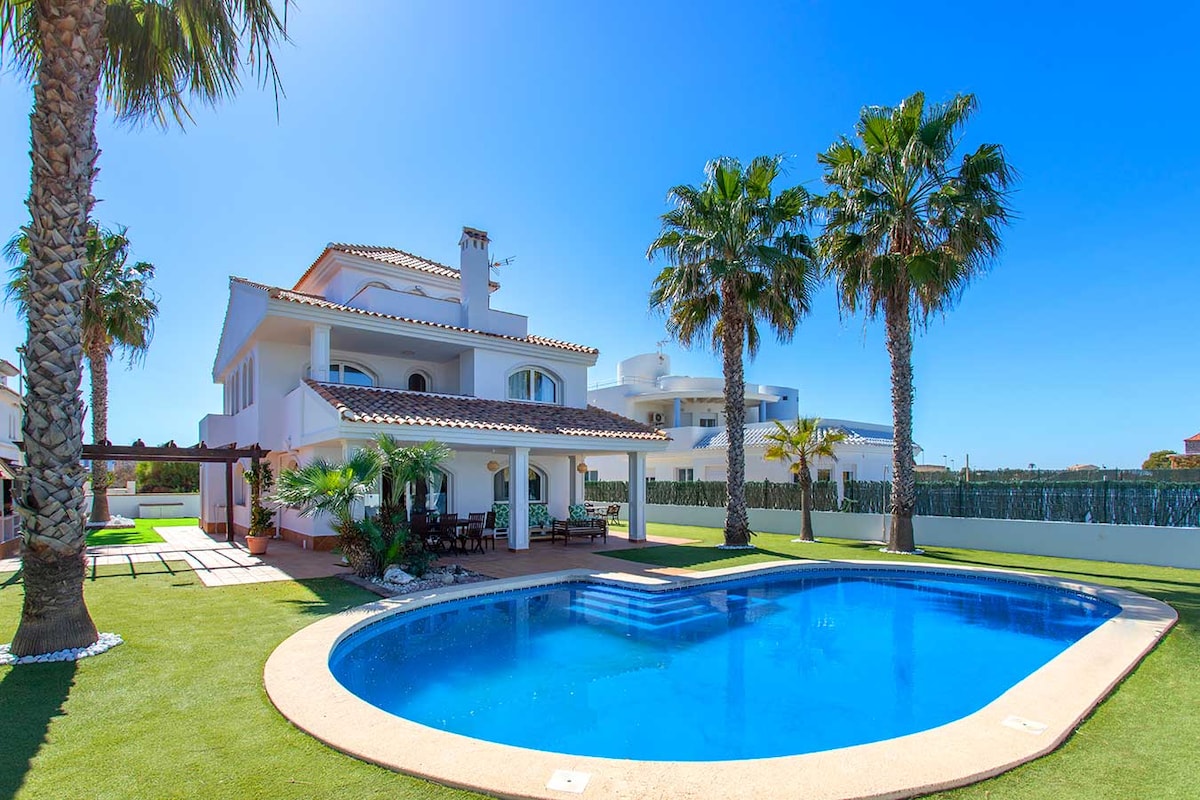 A two-story villa is set under a clear blue sky, featuring a private swimming pool surrounded by lush palm trees. An expansive area of artificial grass presents a welcoming outdoor space, complemented by a shaded patio area adjacent to the house.