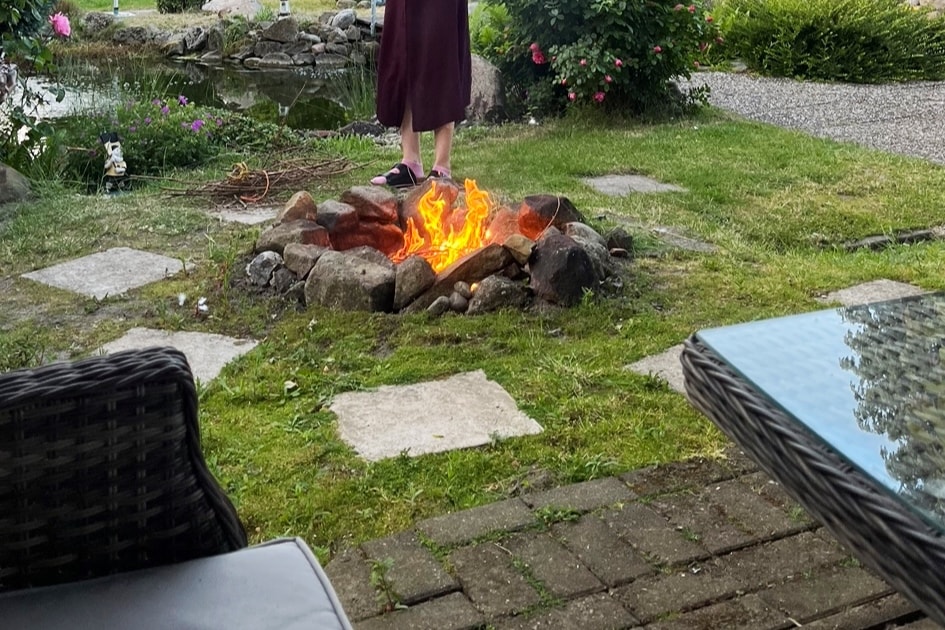 A serene garden setting features a circular stone fire pit with flickering flames, surrounded by lush greenery and colorful flowers. Nearby, a cozy seating area with wicker chairs and a glass table invites relaxation, while a pathway of stepping stones leads through the well-kept lawn.