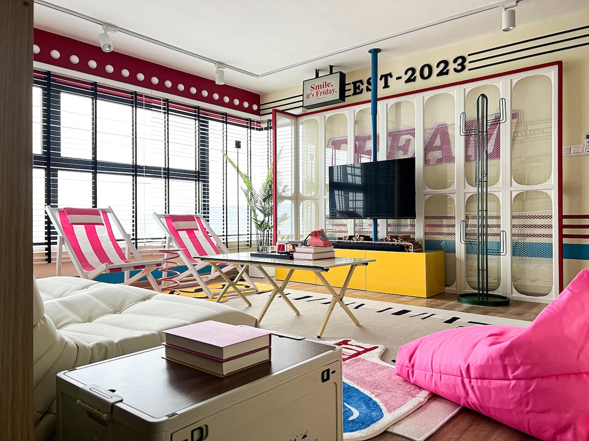 A vibrant living area showcases a retro-inspired design with playful colors. Two lounge chairs with pink and white stripes are positioned for relaxation. A low coffee table is centered, and a television is mounted against a large, patterned wall. Large windows allow natural light to fill the space.