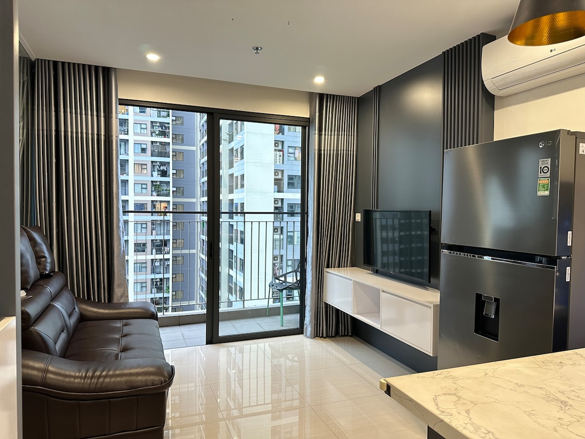 A modern living area is presented, featuring a dark-colored leather sofa positioned near a large glass door that leads to a balcony. A television is mounted on the wall opposite the sofa. Natural light fills the space, enhancing the sleek design of the room.