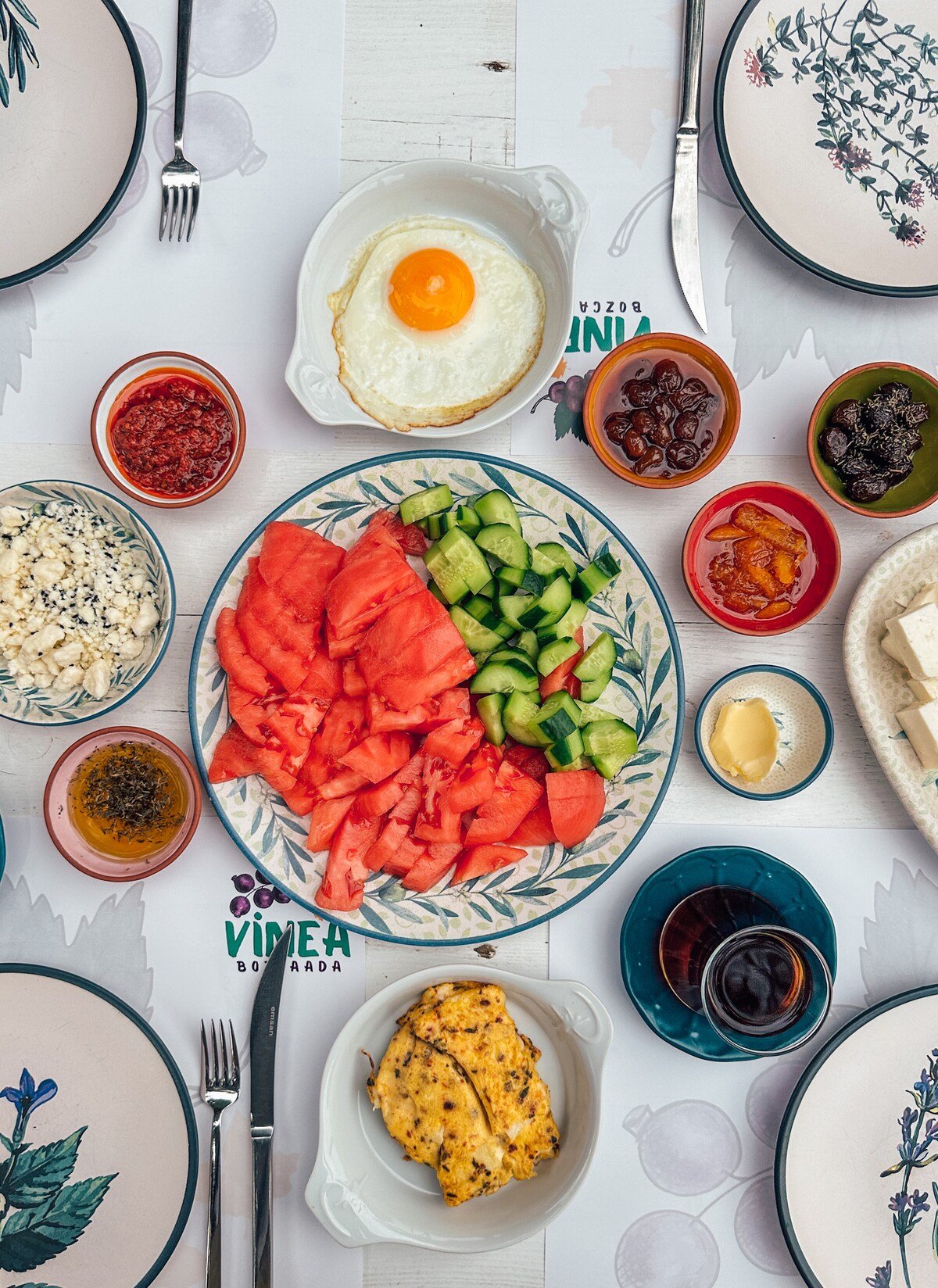 An inviting breakfast spread is displayed on a table, featuring a large plate of watermelon and cucumber slices, alongside a bowl of olives, a fried egg, and various dips. Small servings of cheese and a mixture of jams are also included, creating a colorful and diverse meal.