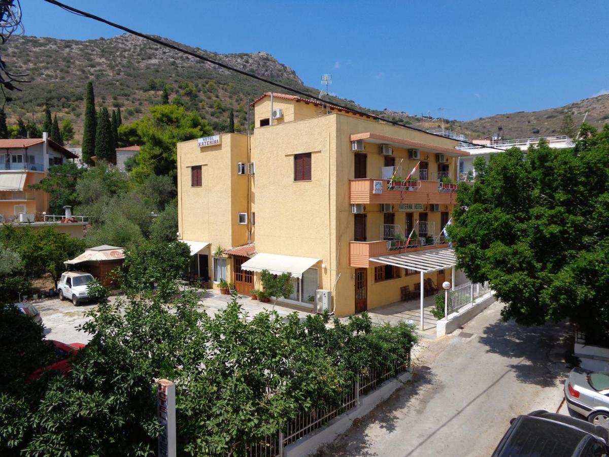 A three-story building with a bright yellow exterior is surrounded by greenery and distant mountains. The structure features several balconies with beige awnings, and a parking area can be seen in the front yard, accommodating multiple vehicles.