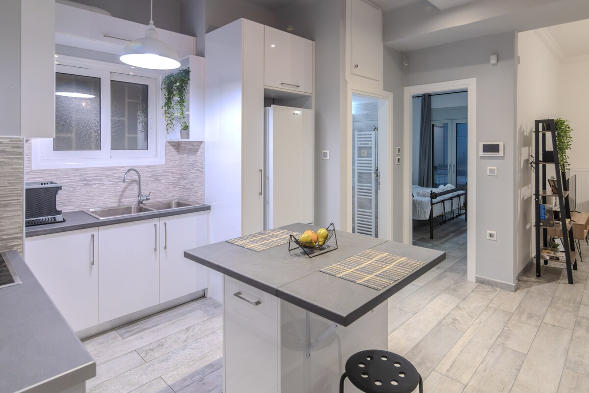 A modern kitchen area features sleek white cabinetry and a large island with seating. Natural light enters through a window above the sink, illuminating the countertop where fresh fruit is displayed. An open layout connects to the living space visible in the background.