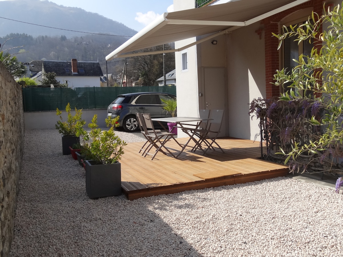 A private terrace features a wooden deck with a small table and chairs, surrounded by potted plants. A vehicle is parked nearby, and a protective awning provides shade. The space is enclosed, offering a sense of privacy amid a tranquil outdoor setting.