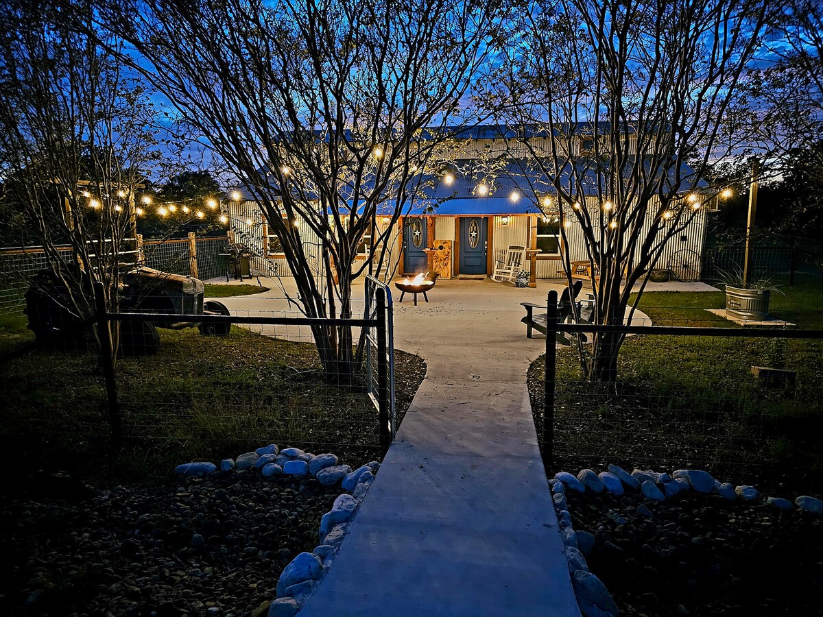 A stone pathway leads to a cozy retreat illuminated by soft string lights at dusk. The entrance features double wooden doors, with a welcoming firepit surrounded by seating positioned on a spacious patio, bordered by greenery and a rustic fence.