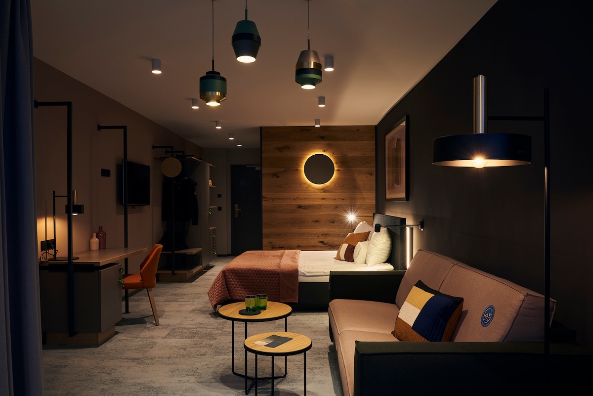 A modern room is furnished with a spacious bed, a sofa, and a stylish workspace. Two small tables are positioned near the sofa, and a warm glow emanates from various pendant lights. A decorative wall adds visual interest to the muted color palette.