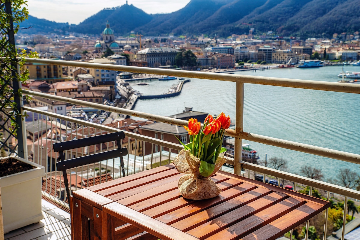 A wooden table is set on a balcony, accompanied by a chair, presenting a bouquet of orange tulips in a natural wrap. A scenic view of the lake and mountains is visible in the background, enhancing the tranquil outdoor space.
