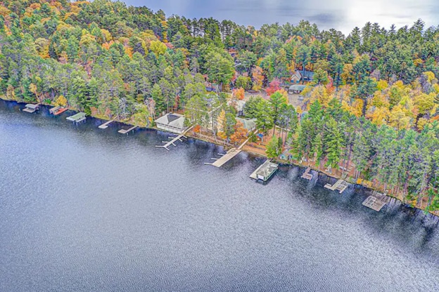 An aerial view captures the expansive landscape of the lodge property, showcasing a private beach and multiple docks along the shoreline. Surrounding trees display autumn colors, and the calm water reflects the scenic beauty of the area, enhancing the natural setting.