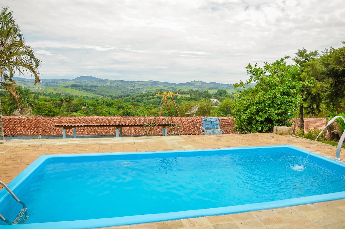 An outdoor area features a blue swimming pool with a gentle water feature, surrounded by a tiled deck. In the background, lush green hills can be seen extending towards an expansive sky, while a swing set is positioned nearby, enhancing the space's appeal for families.