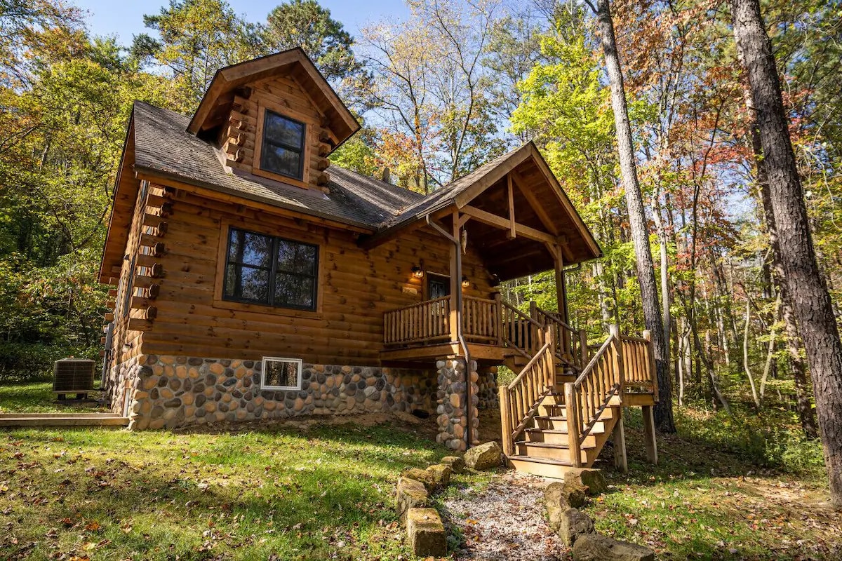 The exterior showcases a traditional log cabin design, featuring a combination of wood and stone. A welcoming front porch leads to the entrance, surrounded by a lush green lawn and trees. Large windows allow natural light to illuminate the interior.