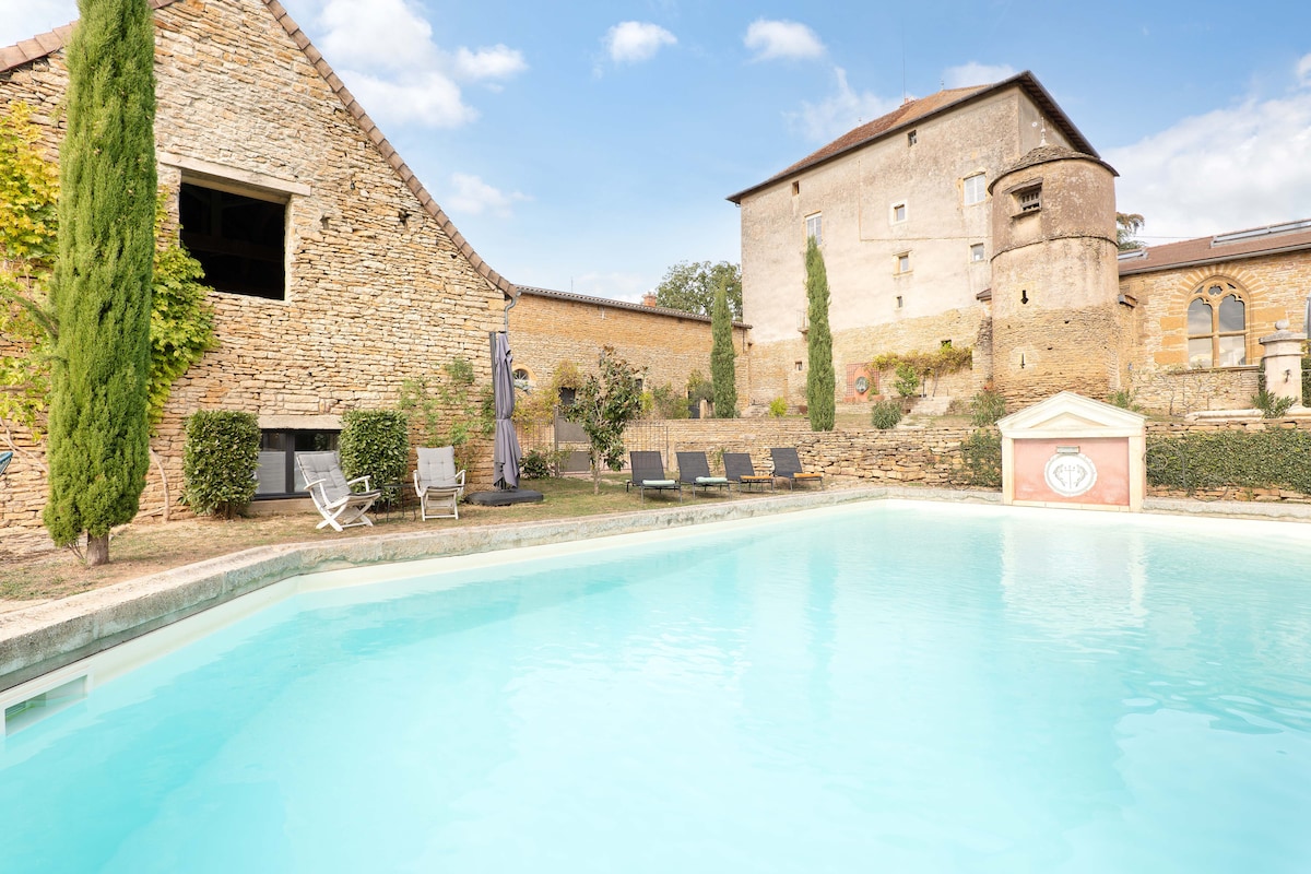 A tranquil swimming pool is set against a backdrop of a historic building, showcasing warm stone walls and lush greenery. Sun loungers are arranged by the poolside, inviting relaxation. The sky is clear, highlighting the serene atmosphere of the outdoor space.