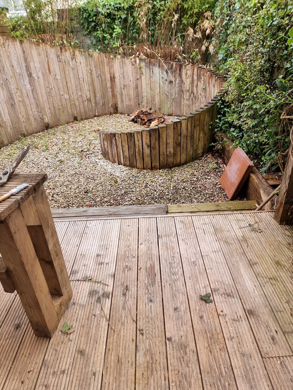 A small enclosed garden features a circular bench made of wood, surrounded by gravel and scattered leaves. A wooden deck extends outward, providing a spacious area for outdoor activities. Greenery lines the perimeter, adding a natural touch to the space.