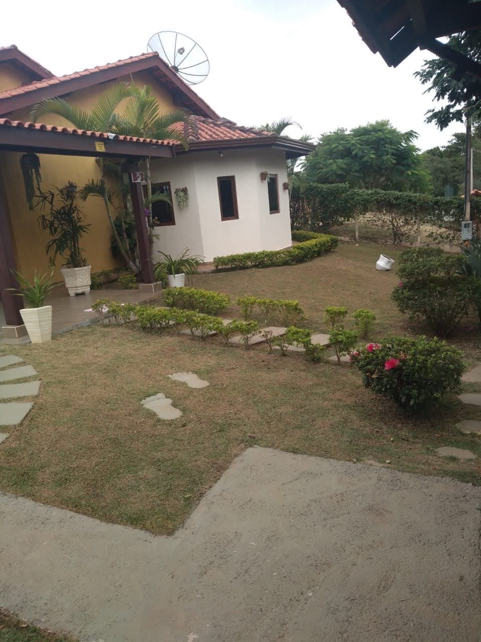 The image shows a well-maintained garden area surrounding a two-tone house. Pathways made of stone lead through the neatly trimmed grass, accompanied by small bushes and flower beds. A satellite dish is visible on the house, with greenery creating a peaceful atmosphere.
