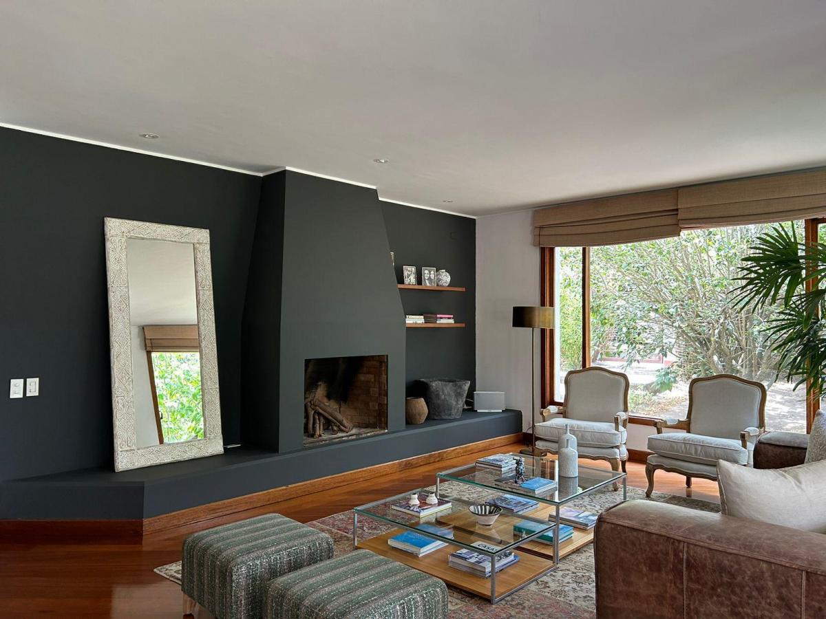 A modern living area features a sleek black accent wall with a stylish mirror and a fireplace. Light from large windows illuminates the space, showcasing two elegant armchairs and a coffee table adorned with books and decor. Natural greenery can be seen outside.