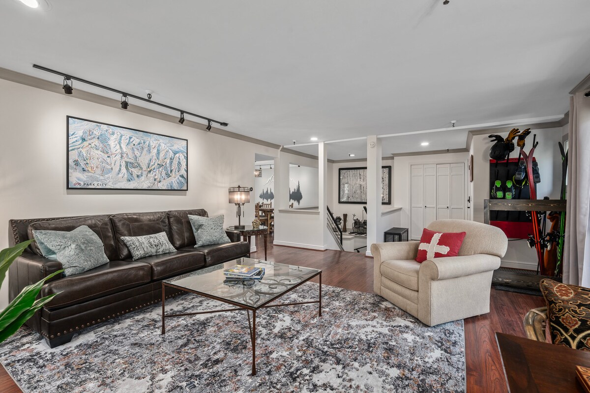 A spacious living area features a comfortable leather couch paired with a cozy armchair. A glass coffee table is centered on a patterned rug, while wall art and soft lighting enhance the room's ambiance. Ski equipment is displayed nearby, highlighting winter sports access.