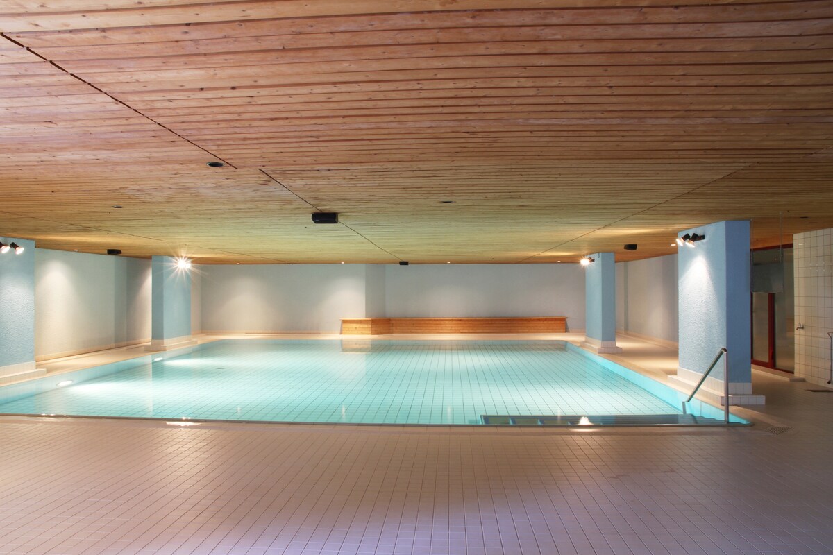 A spacious indoor swimming pool is surrounded by a clean, tiled area. The water appears calm and inviting, with a wooden bench along one side. Soft lighting enhances the tranquil atmosphere, and a ceiling of wooden beams adds a touch of warmth to the space.
