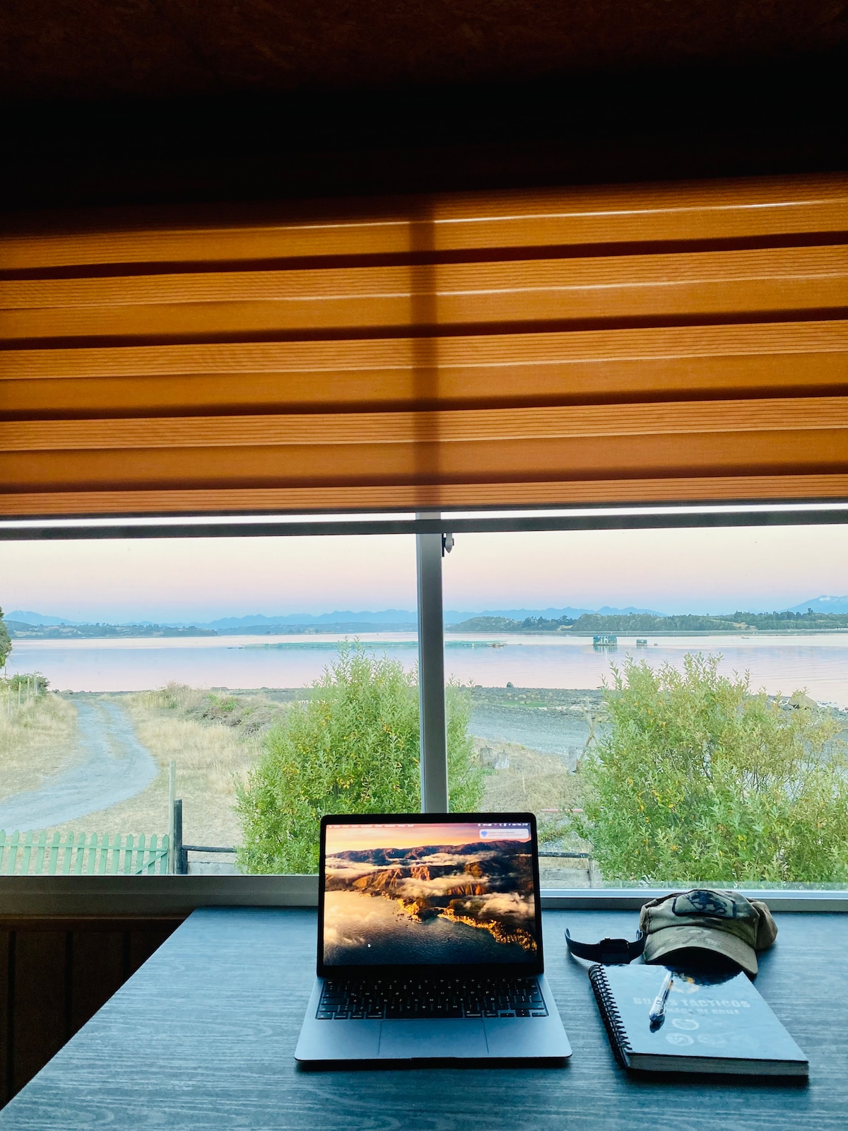 A workspace is set against a window featuring a scenic view of the landscape, including water and greenery. A laptop is open displaying a scenic image. A notebook and a cap are placed on the desk, enhancing the functional and inviting atmosphere of the space.