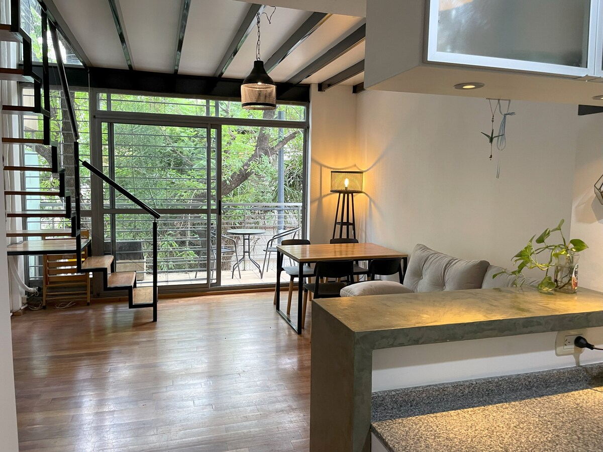 An open living space is illuminated by natural light filtering through large sliding glass doors, which lead to an outdoor area. A cozy sofa is paired with a simple dining table, while a staircase provides access to the upper level. Subtle greenery is visible outside.