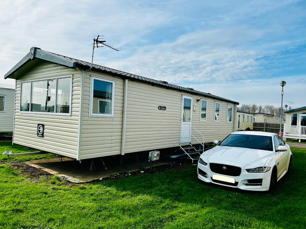 A mobile home is shown with a light exterior, featuring multiple windows along the side. A well-maintained white car is parked adjacent to the home on a grassy area, providing a sense of accessibility. The sky is clear with soft clouds visible above.