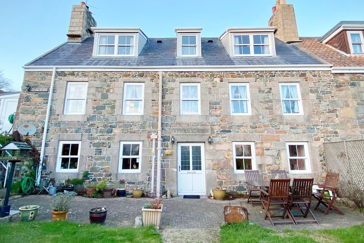 A traditional granite farmhouse with multiple windows and a welcoming entrance. Outdoor seating is arranged in front of the building, surrounded by a garden area. The structure features a classic roofline and stone facade, reflecting its historical architecture.