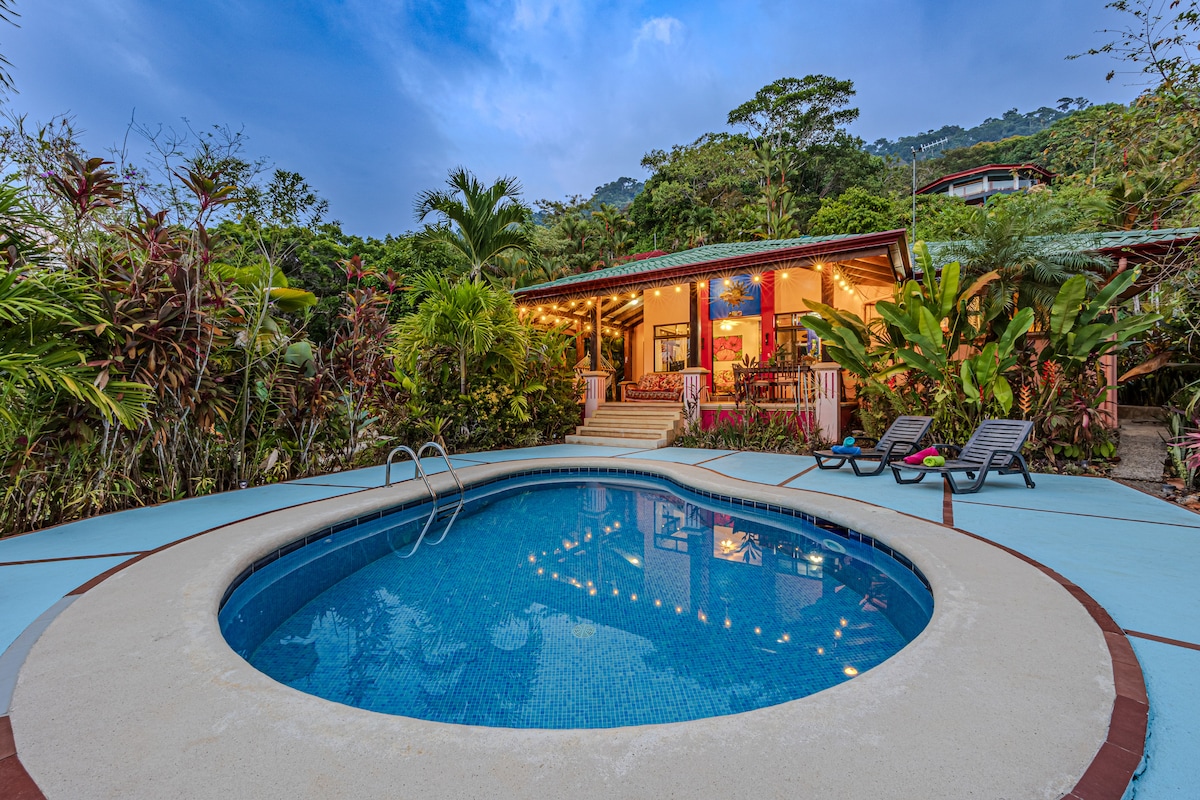 A circular pool is set within a spacious outdoor area, surrounded by tropical foliage and well-maintained landscaping. The home behind features warm lighting and large entryways that connect to the inviting atmosphere, complemented by outdoor seating and charming decor.