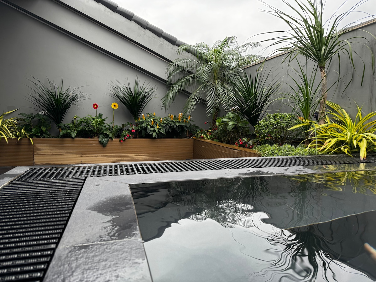 A serene outdoor space features a sleek, dark soaking pool surrounded by lush greenery and colorful flowers. Low wooden planters add a natural element, while tall plants provide privacy. Light reflects gently off the water's surface, creating a calming ambiance.