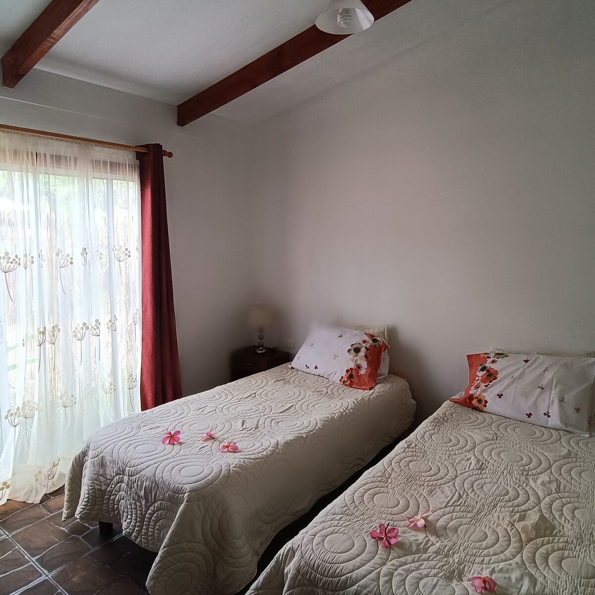 A bright room features two twin beds with decorative floral pillows. Natural light filters through sheer curtains, creating a soft ambiance. The floor is tiled, and a bedside table is positioned next to the beds, complementing the simple décor.