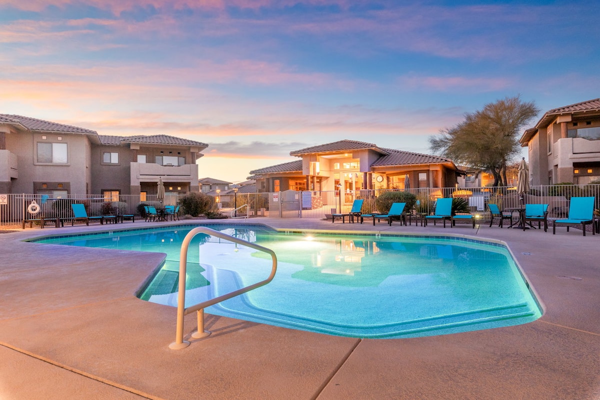 A swimming pool is framed by lounge chairs under a clear sky. The inviting pool features illuminated water reflecting soft sunset hues, while a clubhouse is visible in the background. The surrounding area is designed with pathways and tall desert plants.