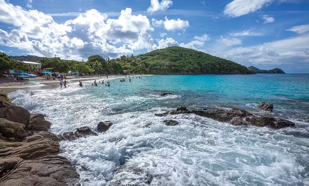 A sandy beach is bordered by smooth rocks, with gentle waves lapping against the shore. Sunbathers and swimmers can be seen enjoying the clear turquoise water, while a lush green hill rises in the background under a partly cloudy sky.