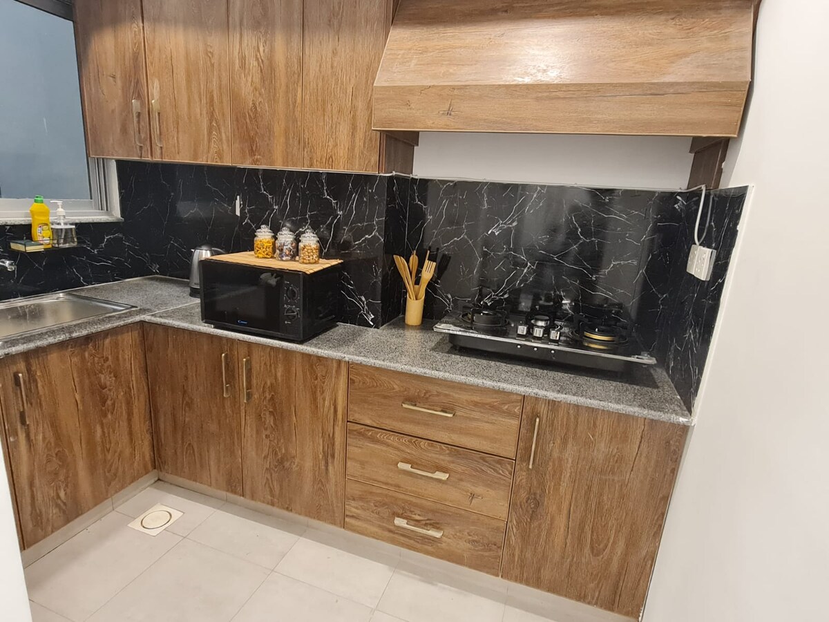 A modern kitchen space features dark marble-like walls contrasted with wooden cabinetry. Essential appliances include a microwave, a stovetop, and utensils displayed in a holder. A countertop provides ample preparation space, while a window ensures natural light enters the area.