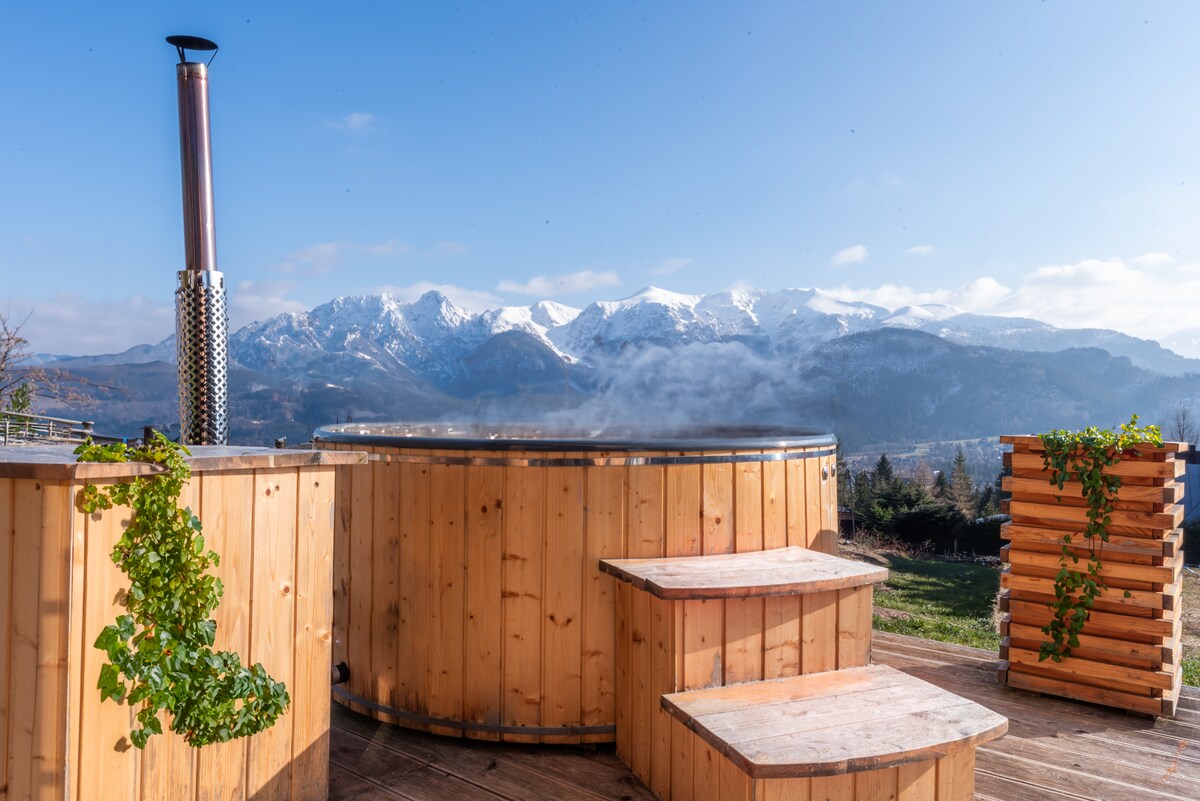 A wooden hot tub sits on a deck, surrounded by steps made of wood and greenery. A scenic view of snow-capped mountains is visible in the background, with clear blue skies above, creating a serene atmosphere.