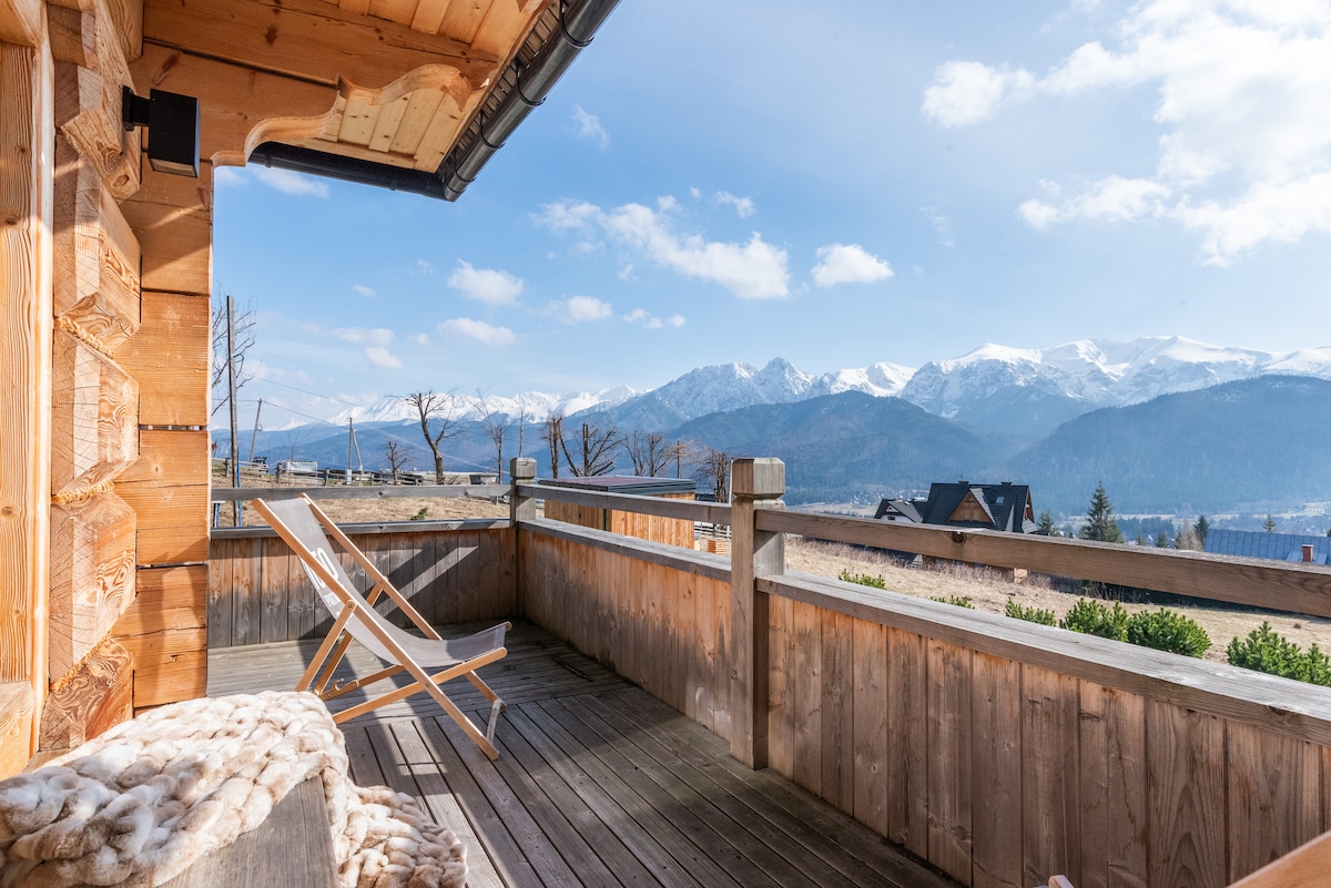 A private balcony features wooden flooring and a lounge chair, offering breathtaking views of the surrounding mountains. The scene is set against a clear blue sky, with distant peaks and greenery enhancing the natural beauty of the location.