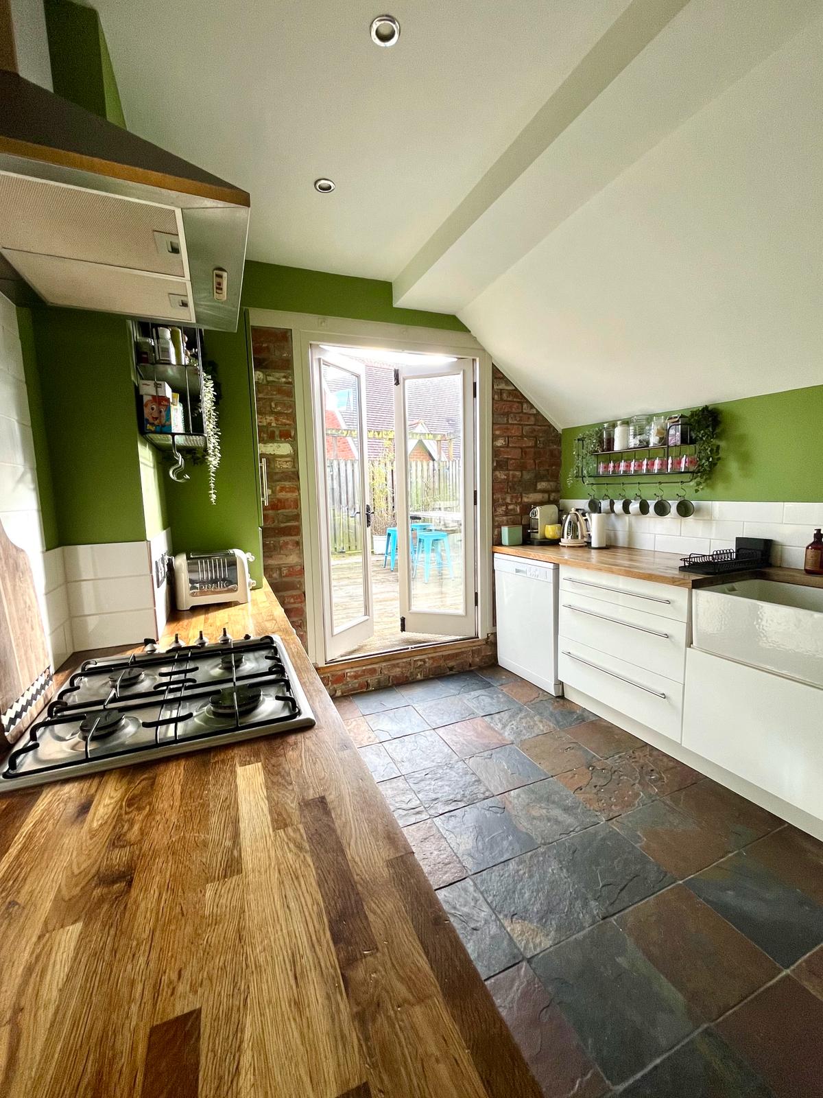 A kitchen with green walls features a combination of modern and rustic elements. A gas stove and wooden countertop are visible, complemented by white cabinetry. Double doors provide access to a private terrace, framed by natural light streaming in from the exterior.