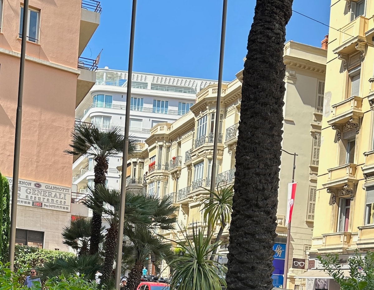 An urban scene captures modern and historical architecture, with a backdrop of classic buildings and contemporary structures. Lush greenery and palm trees can be seen in the foreground, highlighting the vibrant atmosphere of the area. Clear blue skies complete the picturesque view.