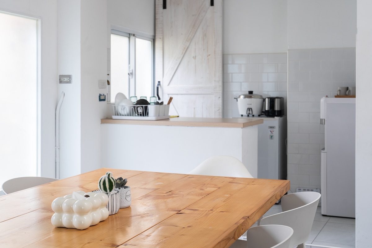 A spacious dining area features a wooden table surrounded by white chairs. In the background, a simple kitchenette is visible, equipped with a refrigerator, coffee maker, and microwave. Natural light filters through large windows, illuminating the calming and minimalistic design.