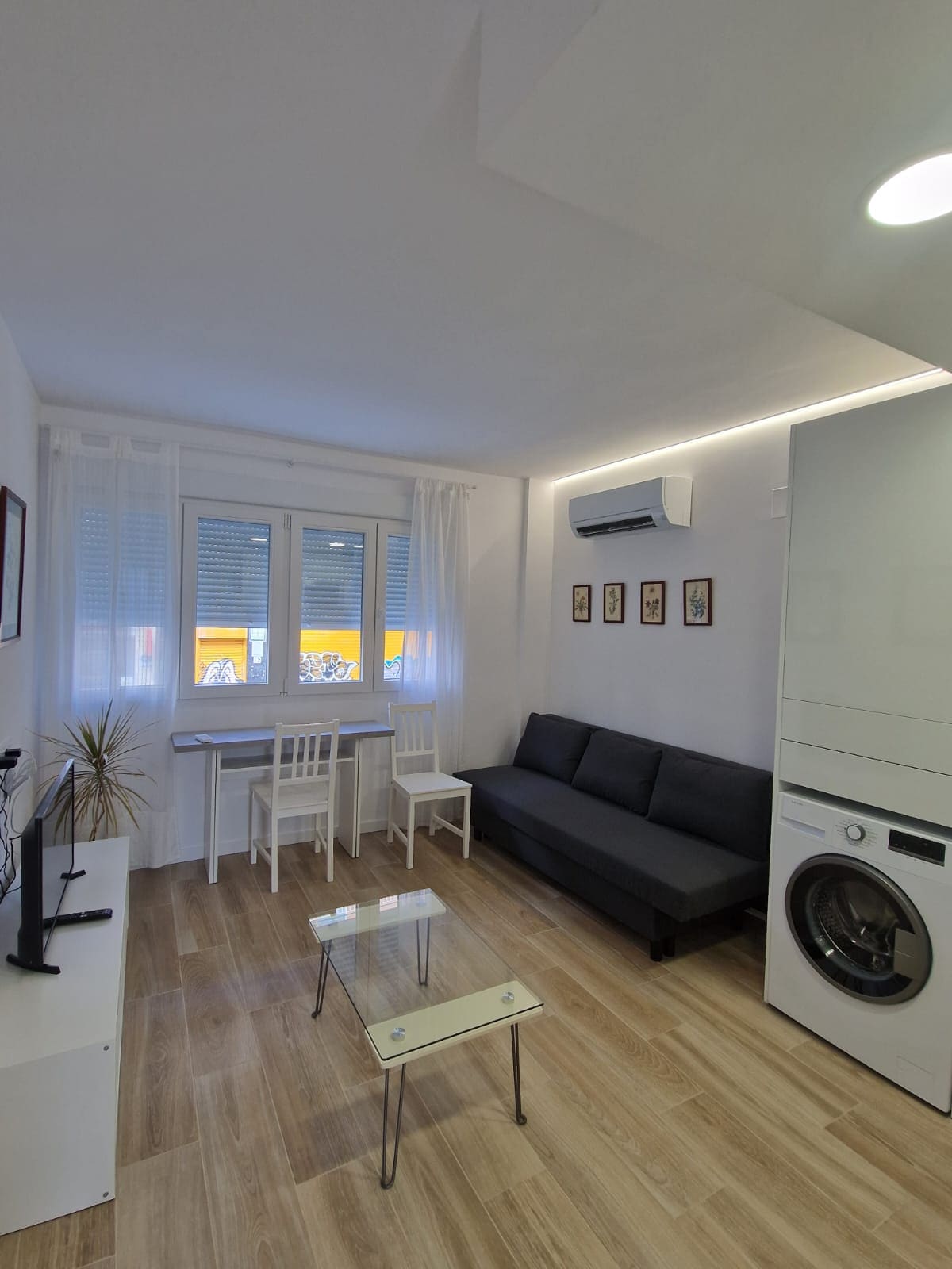 A modern living area is featured, showcasing a dark gray sofa and a glass coffee table. A small dining area with a white table and matching chairs is visible, complemented by large windows that allow natural light to enter, enhancing the bright atmosphere.