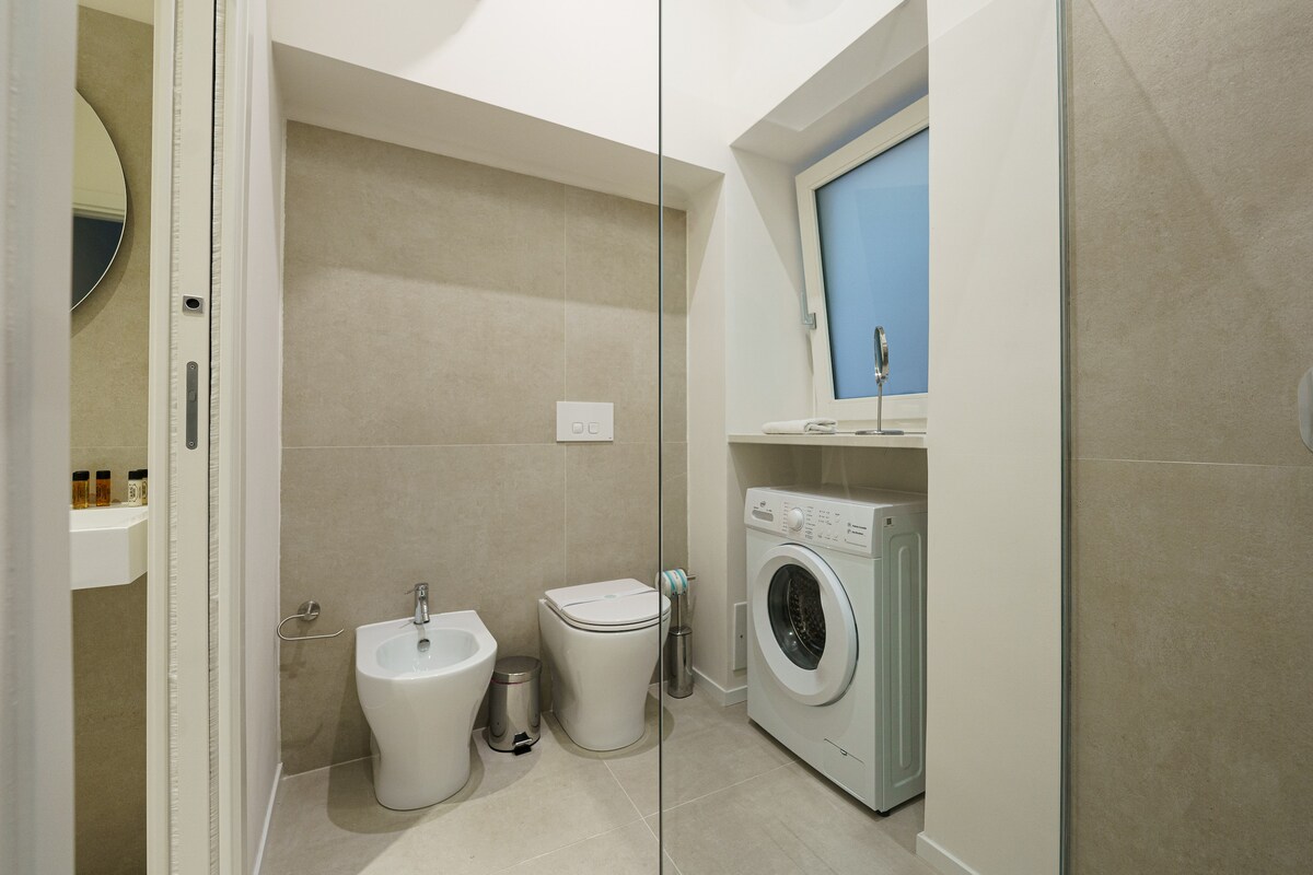 The modern bathroom features a glass-enclosed shower, a toilet, and a bidet. A compact washing machine occupies one corner, while a circular mirror is positioned above the sink. Natural light enters through a small window, illuminating the neutral-colored tiles and fixtures.