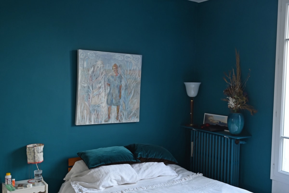 A cozy bedroom features a large bed with white linens and two teal pillows. A painting of a figure in a blue outfit is mounted on a textured turquoise wall. A bedside table holds personal items, while a blue vase with dried flowers adds a touch of decor.
